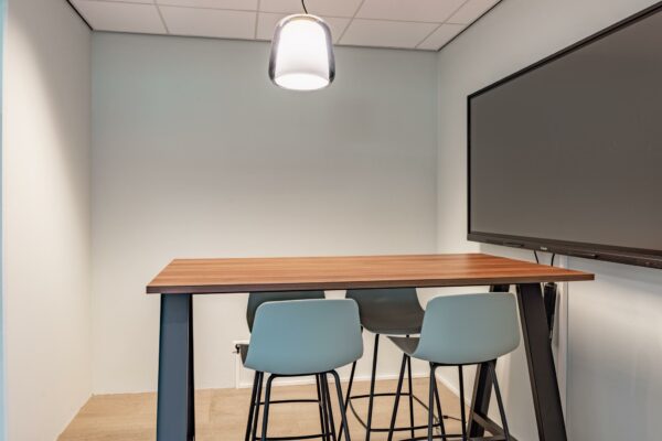 Moderne vergaderruimte met hoge tafel, vier barkrukken, hanglamp en een groot digitaal scherm aan de muur.